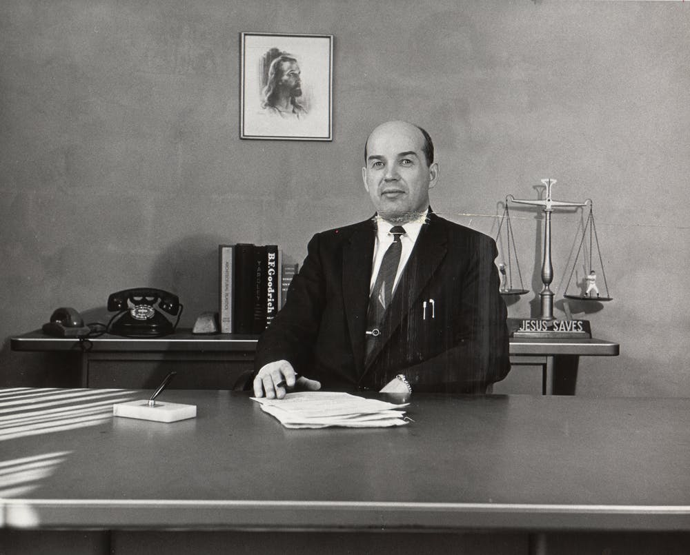 Stanley posing behind his office desk with documents in front of him. A portrait of Jesus hangs on the wall behind him