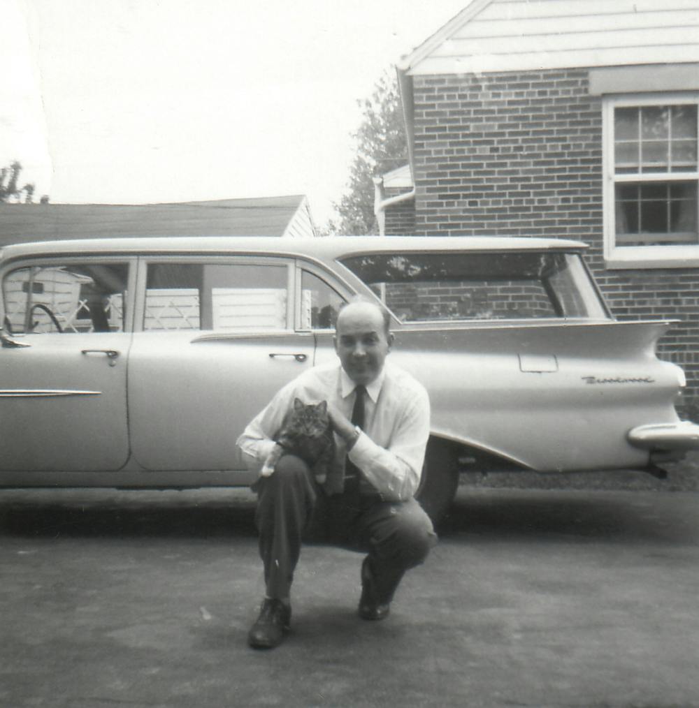 Stanley squatting in front of a car while holding a cat